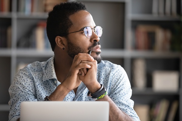 Homme afro-américain pensif portant des lunettes, distrait par son travail sur ordinateur, regardant au loin, pensant ou réfléchissant
