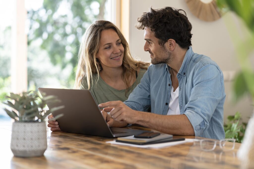 Un jeune couple se regarde en souriant devant un écran d’ordinateur.
