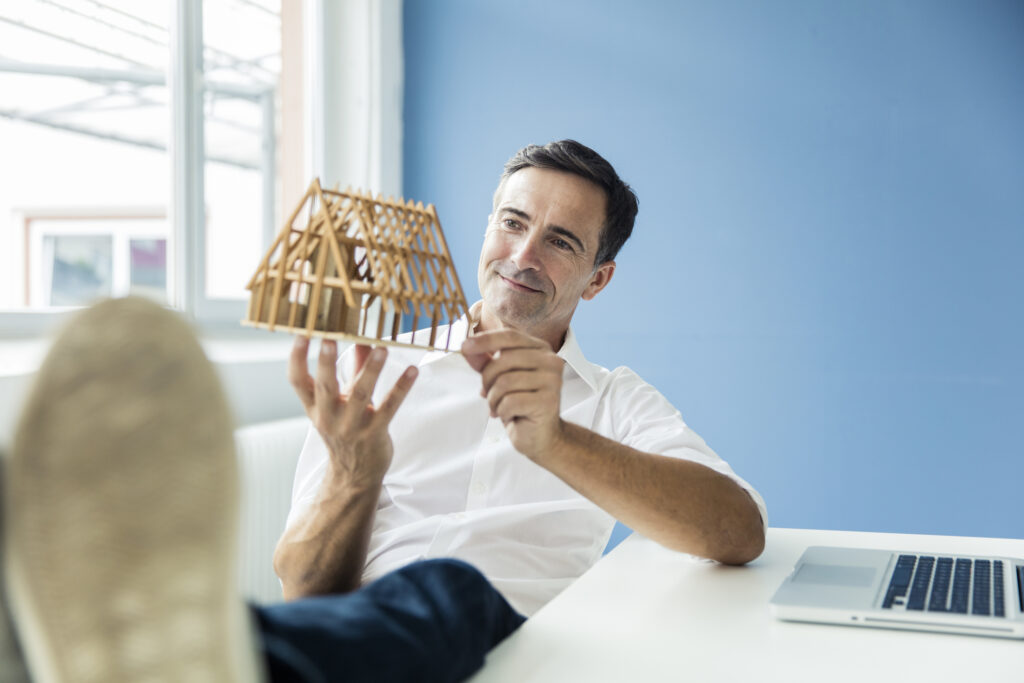 ​​Un homme, les pieds posés sur son bureau. Il tient entre ses mains une maison à l’état de maquette.​
