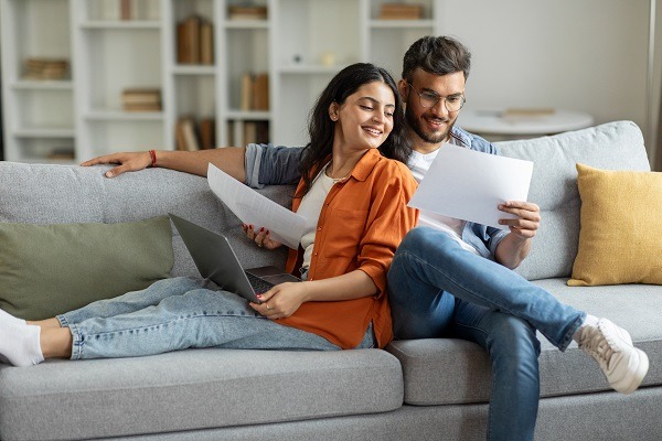 Couple indien heureux vérifiant des documents, conjoints lisant un accord d'assurance ou un certificat de propriété, mari et femme assis sur un canapé dans un salon
