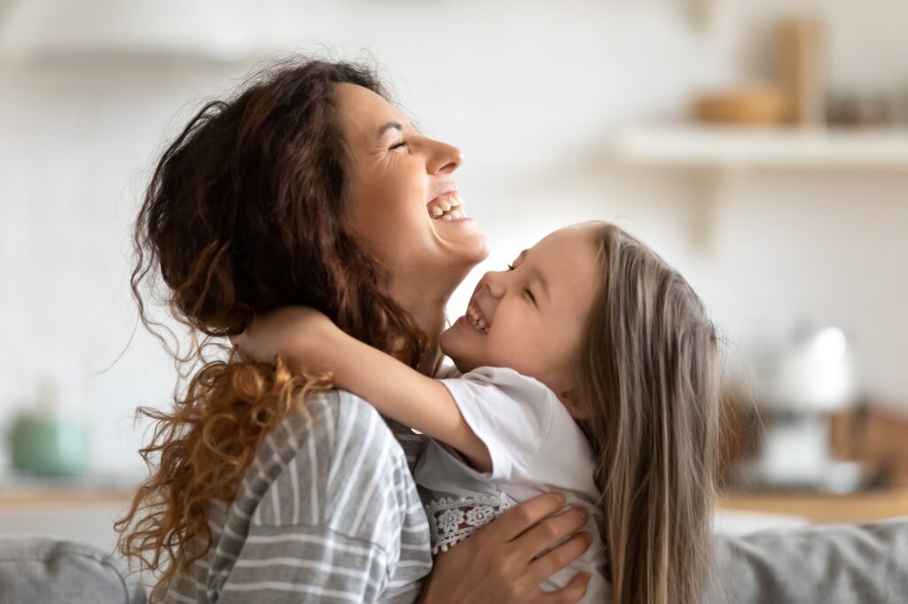 Vue latérale en gros plan d'une jeune mère et de sa fille souriantes qui se serrent dans les bras et rient, profitant d'un moment de tendresse, d'une maman heureuse et d'une adorable petite fille d'âge préscolaire qui se câlinent, s'amusant ensemble à la maison