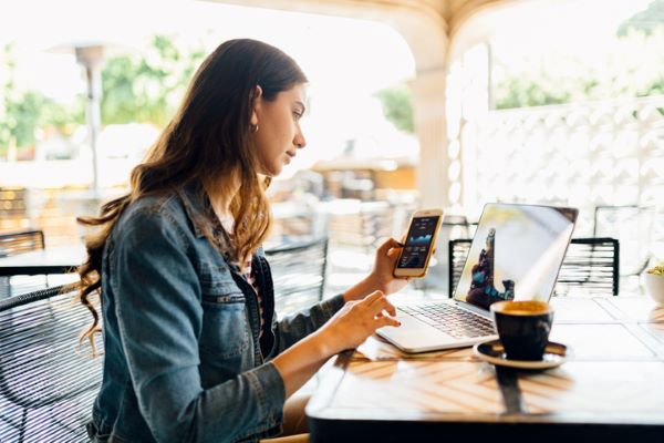 Une jeune femme assise à une terrasse couverte consulte son téléphone mobile en regardant l’écran de son ordinateur portable posé sur une table devant elle avec une tasse de café à côté