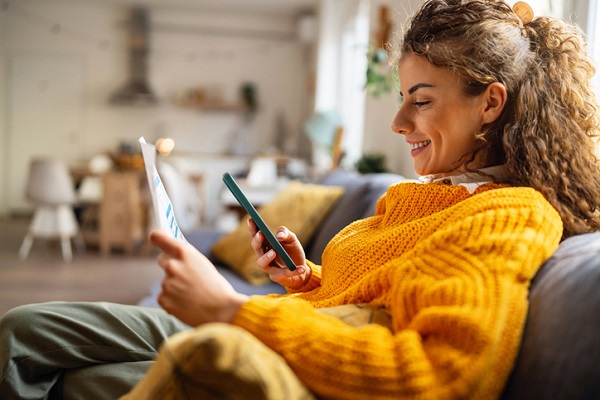 Une jeune femme assise sur son canapé, tient son smartphone d’une main et regarde un document qu’elle tient de l’autre main
