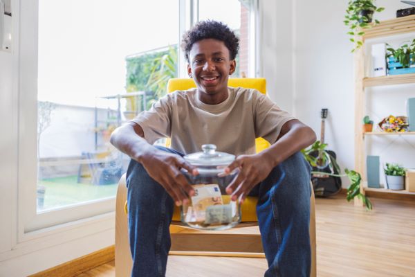 Un adolescent souriant tient entre ses mains un pot transparent tirelire contenant des billets et pièces, assis sur une chaise à la maison, regarde le photographe de face.